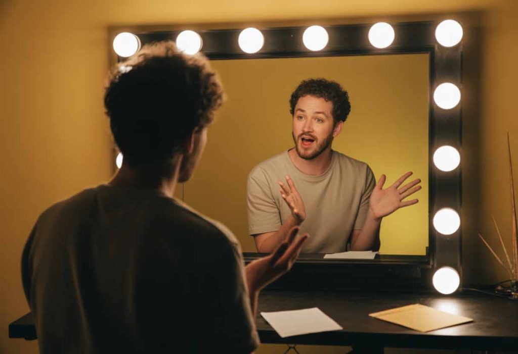 Comedian practicing punchlines with expressive gestures in front of a mirror.